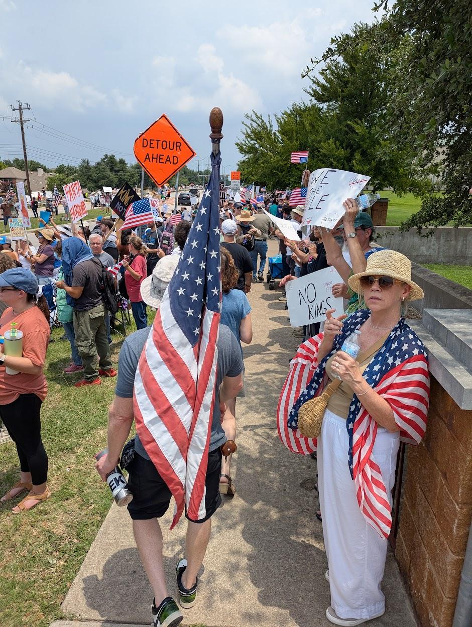 People walk down a sidewalk lined with protesters. A number of the protesters are carrying or waving US flags.