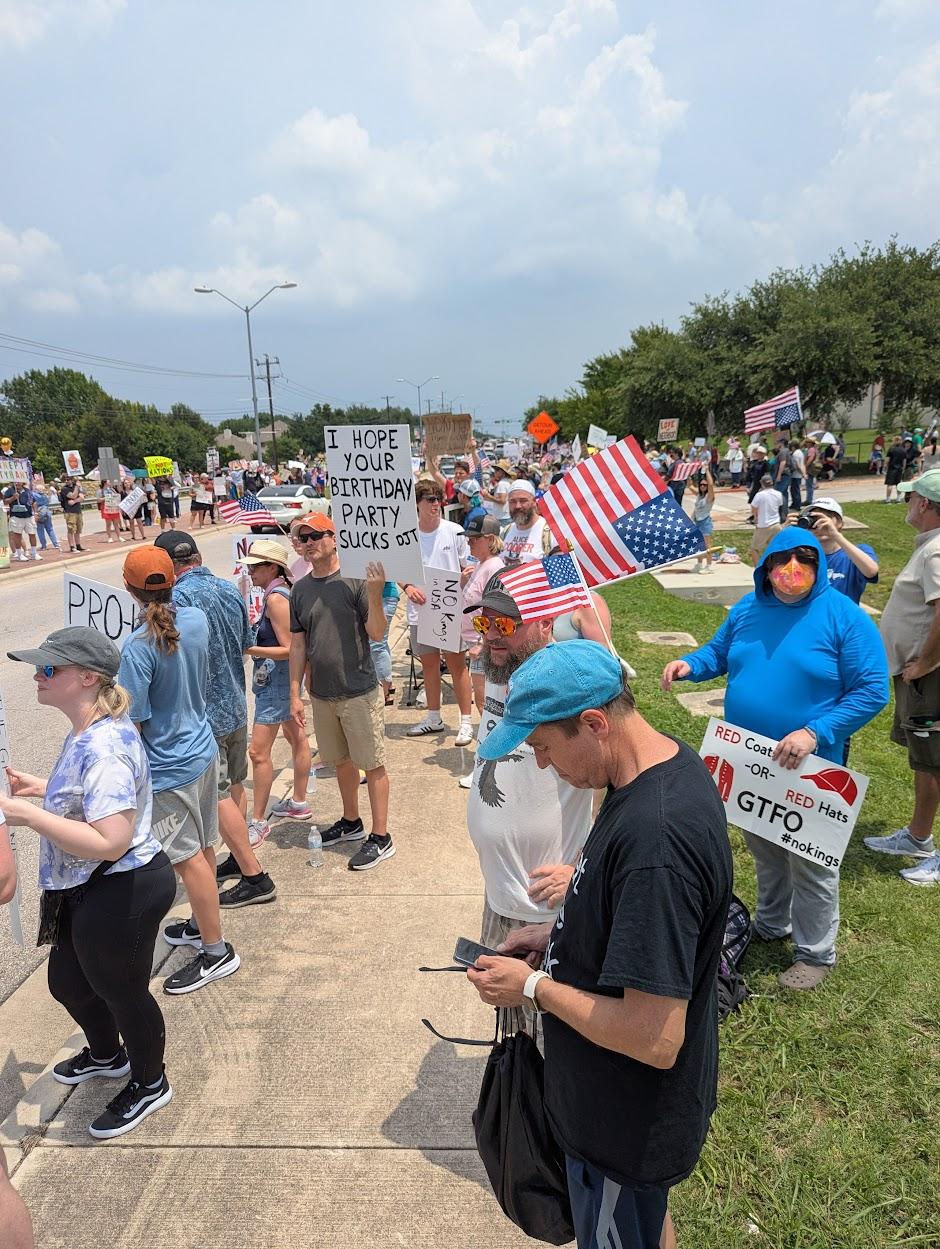 Looking one direction down the street. A number of protesters line the street on both sides and in the median. One sign reads, "I hope your birthday party sucks, DJT." Another reads, "Red Coats or Red Hats, GTFO. #NoKings"