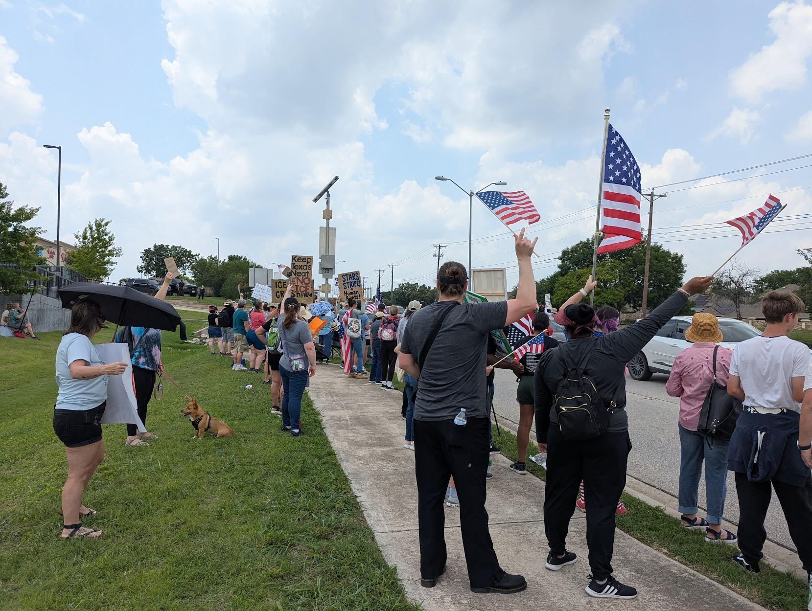 Looking the other direction down the street. A line of protesters with waving US flags and posters, and a cute dog. One sign reads, "Keep Texas neat. No ICE." Another reads, "Stars and Stripes and Human Rights."