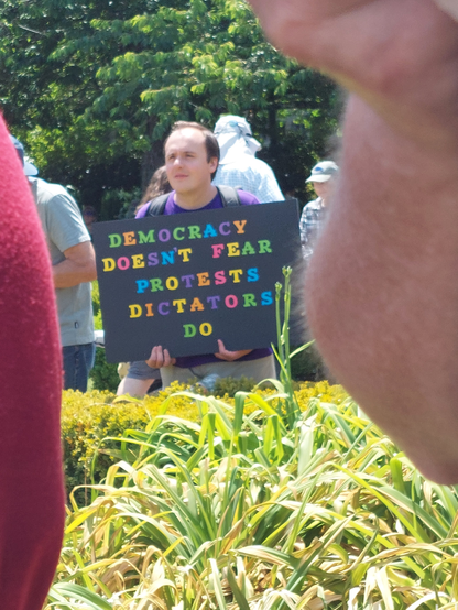 A man seen in the distance with a colorful sign saying, "Democracy doesn't fear protests dictators do"