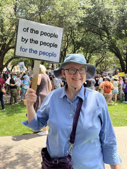 A woman in a blue blouse and a floppy sun hat holds a sign above her head that says "of the people, by the people, for the people.