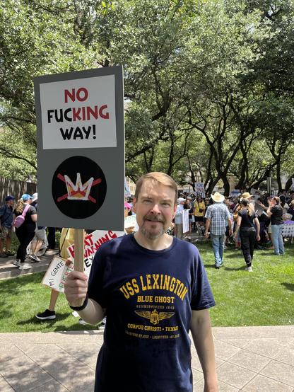A man in a U.S.S. Lexington t-shirt holds a sign above his head that says No Fucking Way with the letters NO and KING in bright red and, with a graphic of the "no kings" logo.