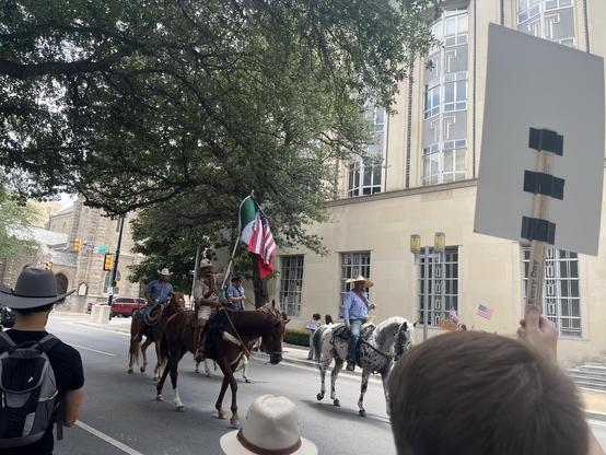 Four cowboys carrying Mexican and American flags ride horses down the street past a crowns of people watching from the sidewalk.