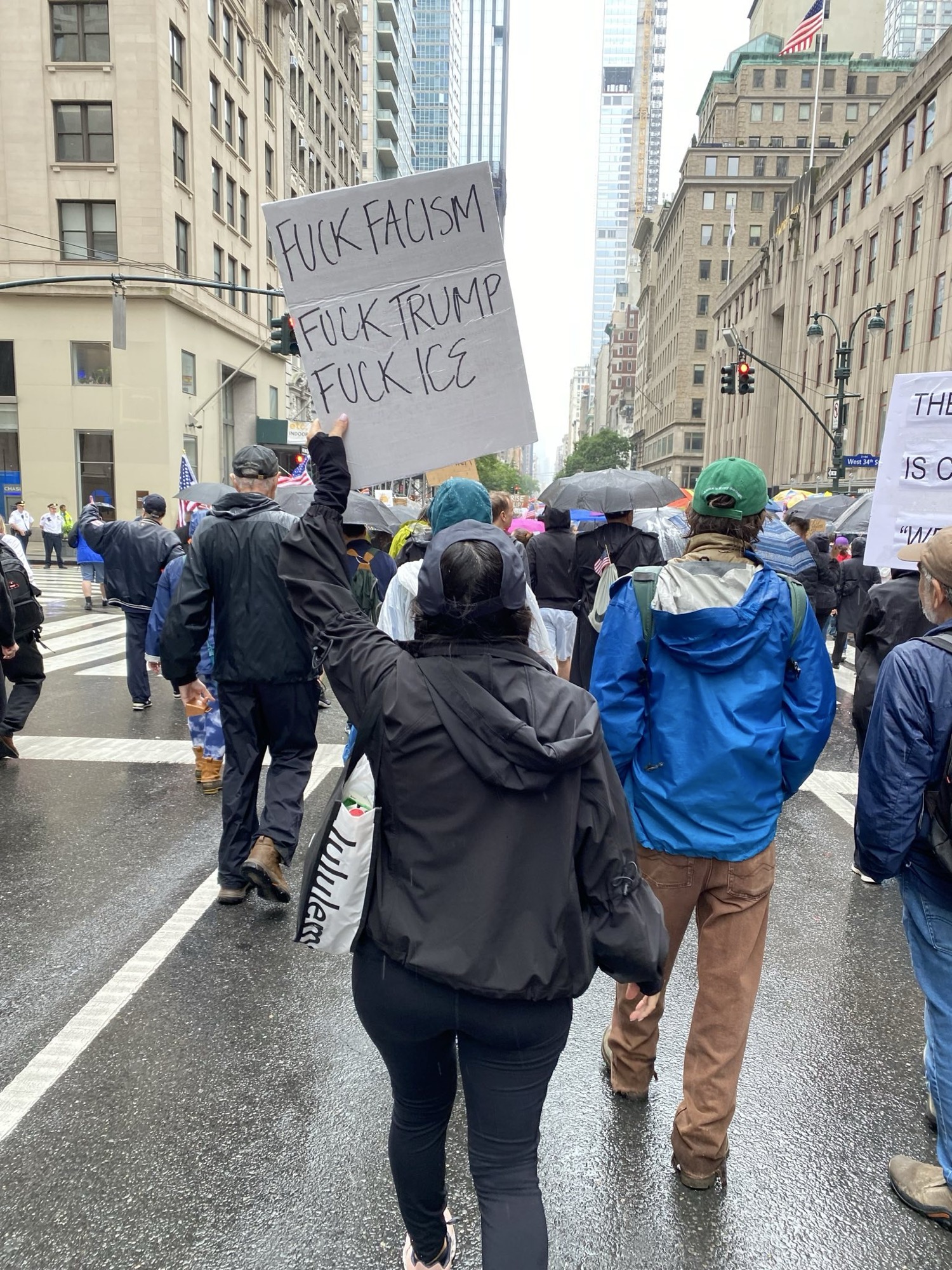 A photo of a person’s back during a protest. They are holding a sign that says “Fuck fascism Fuck Trump Fuck ICE”