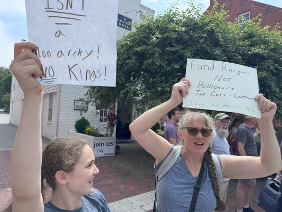 Woman and young girl holding signs.  One reads "Fund Rangers not billionaire tax cuts"  other is cut off but reads "America Isn't a Monarchy: No Kings"