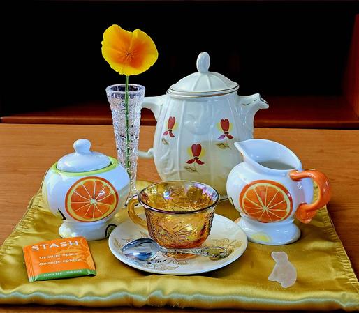 Sunny, warm tea tableau on gold satin with marigold Carnival glass, a crystal vase holding a fresh California poppy, a Sadler teapot, and sugar and creamer with a pattern of orange slices. The spoon has a tiny California brown bear symbol, and there is a frosted glass bear. The teabis Orange Spice black tea from Stash.