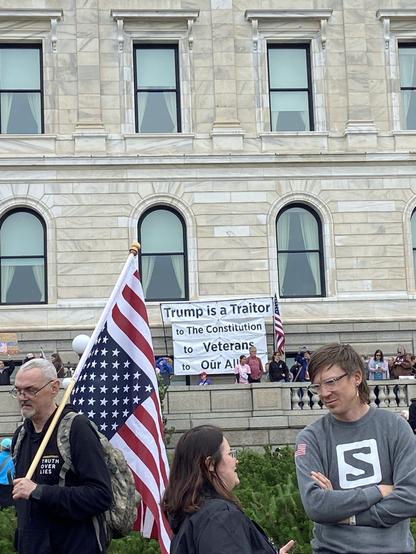 Demonstrators in foreground include a person carrying a pole-mounted, upside-down United States flag. A banner in the background reads:  Trump is a Traitor to The Constitution to Veterans to Our Allies