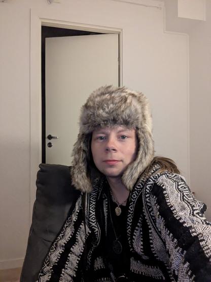 A person wearing a fur hat and patterned poncho sits calmly on a dark couch in a dimly lit urban home, giving an eccentric, introspective impression.