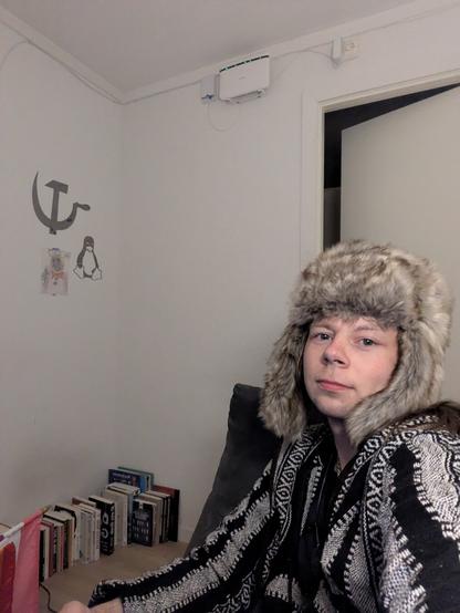 A young adult in a fur hat and patterned poncho sits in a modest living space; background features a stack of books, a wall-mounted router, and a wall drawing of a hammer and sickle, robot, and penguin.