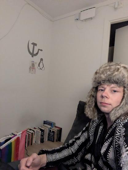 A young adult in a fur hat and patterned poncho sits in a modest living space; background features a stack of books, a wall-mounted router, and a wall drawing of a hammer and sickle, robot, and penguin.