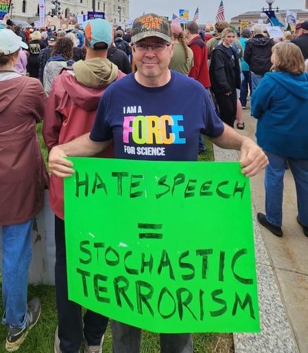 A man wears a hat that says CEASE FIRE in blaze orange. 

He dons a T-shirt that reads I AM A FORCE FOR SCIENCE 

He holds a sign that reads: НАТЕ SPEECH = STOCHASTIC TERRORISM