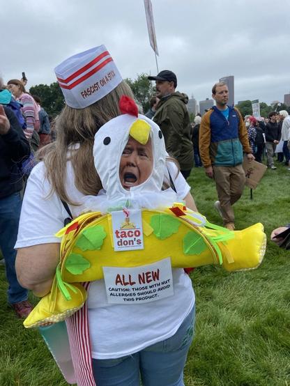 Rear view of protesters/artist. She dons a food-service hat labeled FASCIST ON A STICK, and has a likeness of a large Taco strapped to her back labeled “taco don’s”