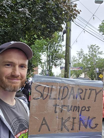 Man holding a cardboard sign with the words "SOLIDARITY trumps A KING". In the background you can glimpse other protesters, trees, and a telephone pole. It is a wet and cloudy day.