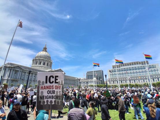 A crowd in front of San Francisco City Hall, framed with pride flags, and a sign saying "ICE hands off our neighbors"
