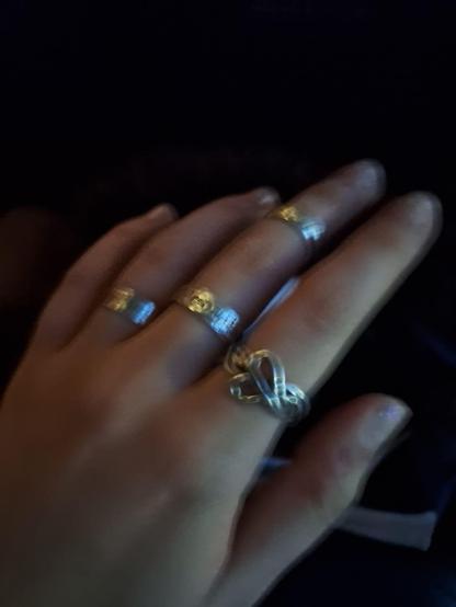 The image shows a close-up of a hand with four rings on the fingers. The hand is positioned with the palm facing upwards, and the fingers are slightly spread apart. The rings are of different styles and materials. Three of the rings are gold-colored with a simple band design, and each has a small, circular, embossed detail in the center. The fourth ring is silver and features a more intricate design with a twisted band and a knot-like structure. The nails are short and have a natural, unpainted appearance. The background is dark, which makes the rings stand out. The lighting is low, creating a soft glow around the rings, particularly highlighting the gold ones.

Provided by @altbot