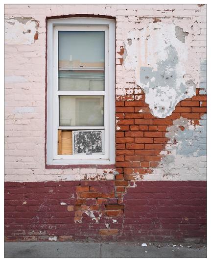 Vertical image featuring a section of a weathered building wall with a mixture of peeled paint and exposed bricks. On the left, a white-framed window, partially shadowed, includes a small poster with text visible inside it. The surrounding wall has layers of faded red and white paint, with significant patches of exposed reddish brick and crumbling mortar. A narrow sidestreet is partially visible at the bottom.