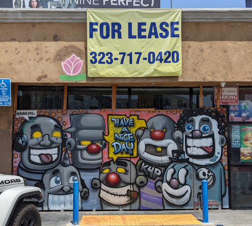 For Lease sign over a boarded up storefront with a great gang of faces painted on the boards