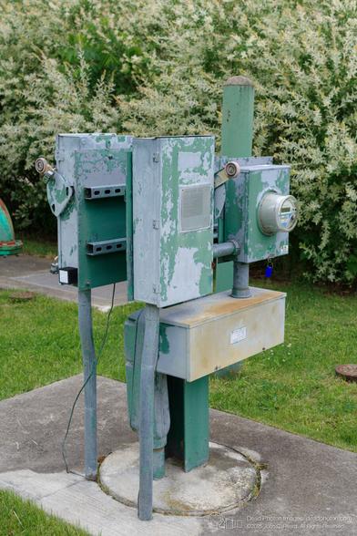 A utility structure stands on a concrete pad surrounded by grass and a flowering bush. The structure is composed of several metal boxes and pipes, painted in shades of green and gray, with significant paint chipping and rust. The boxes appear to house electrical or metering equipment. One box has a visible meter with a digital display. The structure is likely used for monitoring or controlling utilities such as water or electricity. The background features a vibrant green lawn and a large bush with white and green foliage, providing a natural contrast to the industrial appearance of the utility structure.