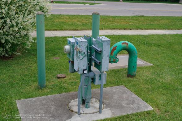 Green utility equipment on a concrete pad in a grThis image shows a utility installation located on a grassy area next to a road. The installation consists of several components, all painted in a faded green color. Two cylindrical posts stand vertically, flanking the main structure. The main structure is a collection of metal boxes and pipes, appearing somewhat weathered with peeling paint and rust. A curved pipe, shaped like an upside-down U, is also part of the installation. The entire structure sits on a square concrete pad. In the background, a road is visible, and a bush with white flowers is to the left of the installation. The overall impression is of a functional, but slightly neglected, piece of infrastructure in a suburban or residential setting.
assy area. The equipment includes pipes, boxes, and poles.
