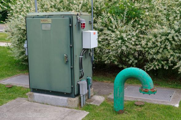 A utility box and associated infrastructure are situated on a grassy area next to a road. The box, painted a dull green, is labeled AGGRESSIVE SYSTEMS INC. and has a red warning light on its side. Various conduits and electrical boxes are connected to it. A large, curved, turquoise pipe with peeling paint rises from the ground nearby. The pipe is connected to a metal plate set into a concrete base, which is likely a manhole cover. The scene suggests an underground utility system, possibly related to water or sewage. The surrounding vegetation includes a bush with white-tipped leaves, adding a touch of natural color to the otherwise industrial setting. The overall impression is one of functional infrastructure, slightly weathered and integrated into the landscape.