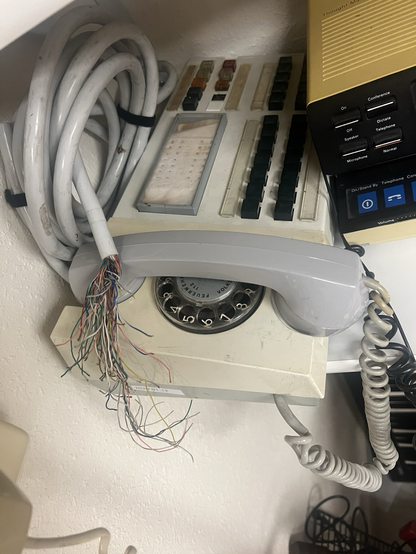 white telephone with rotary dial under the handset. many unlabelled black buttons the writing has long since faded. there is a thick bundle of twisted pair wires still attached to the phone