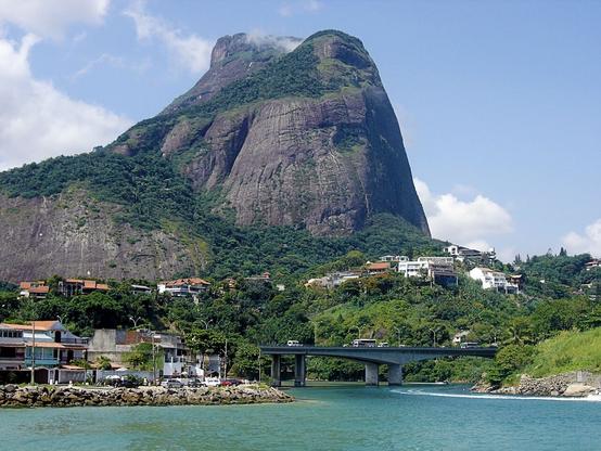 A scenic view featuring a towering mountain (Pão de Açucar) with lush greenery, overlooking a body of water. In the foreground, there are residential buildings and a bridge crossing over the water. The sky is partly cloudy, adding to the serene landscape.