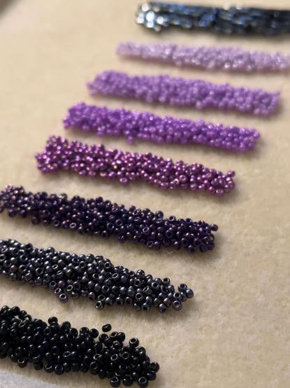 A line of seed bead piles in shades of purple, grey, and black, on a cream colored bead mat