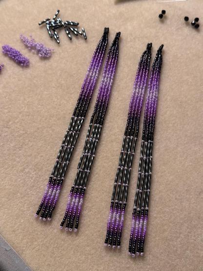 Two pairs of long beaded dangle earrings in shades of purple, gray, and black, on a bead mat. There are piles of bicone beads, bugle beads, and seed beads in the same colors around the bead mat.