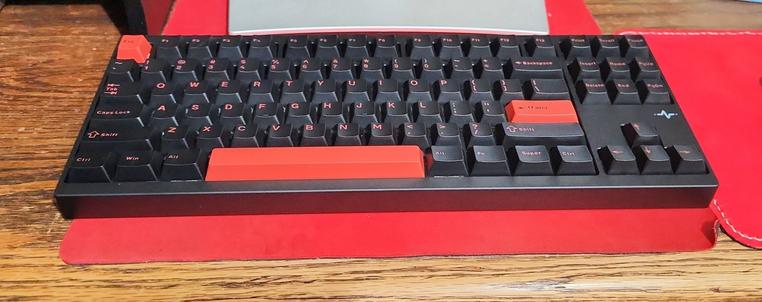 A snazzy black and red keyboard sits on a red desk mat, all on a wooden desk. A matching red mousepad is off to the right side.