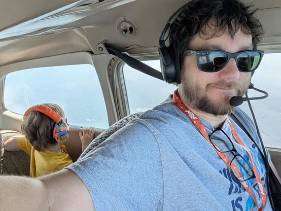 A man flying an aircraft taking a selfie that shows a child in the back of the aircraft peering out at the wonders below.