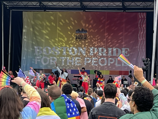 A somewhat distant photo from the crowd. The crowd is waving their hands. As is everyone on stage. Big Freedia is wearing a rainbow skirt and a denim top. There’s a large banner behind the performers reading Boston Pride for the People. It’s rainbow colored.