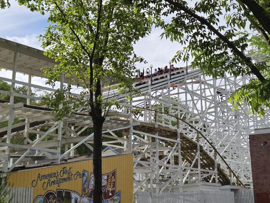 Detail of the white-painted lift hill structure of a classic wooden roller coaster. It's a partly sunny sky. There is a coaster train ascending the lift, full of riders some of whom already have their arms in the air, framed by the leafy branches of trees in the foreground. Just in front of the lift is the very end of the roller coaster's track, rising into the covered brake run, with some of its structure in white and some in unpainted wood. The brake run is supported by a building with a slanted roof at bottom of frame, with a sign in vintage postcard style identifying Lake Compounce as "America's First Amusement Park."