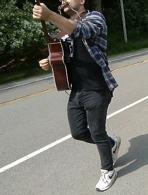 A musician at the Irondequoit NY No Kings rally on June 14 2025. The picture shows a man walking on the shoulder of a road, with trees visible behind him across the street. The man is playing "This Land Is Your Land" on an acoustic guitar, accompanying himself on a harmonica held up to his mouth by a holder. He is wearing dark jeans, a dark Tshirt, sneakers, and an unbuttoned plaid flannel shirt. The mouth and lower part of his face are visible, but the upper part of his face is cut off at the top of the photo.