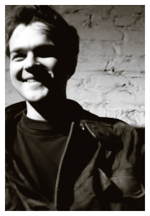 night. lit by a nearby bare bulb. 20s man with shortish dark hair wears a dark tee and dark suede jacket and with broad smile. the background is very old brick wall, painted white.