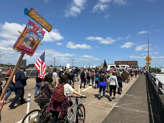 Large crowd marching along the east side of the Hawthorne Bridge. Foreground sign says "America has a problem. No kings" with a painted image of Beyoncé in Cowboy Carter outfit and "Bey" underneath