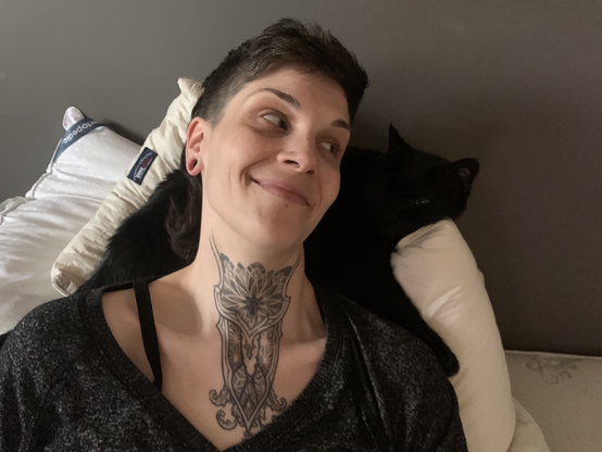 Woman resting her head on a black cat laying on a pillow, the woman smiling looking towards the cat. The cat is bruce, who is being fundraised for his vet bills.
