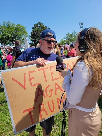 A man holding a "Veterans Against Trump" sign is being interviewed by a journalist at an outdoor gathering.