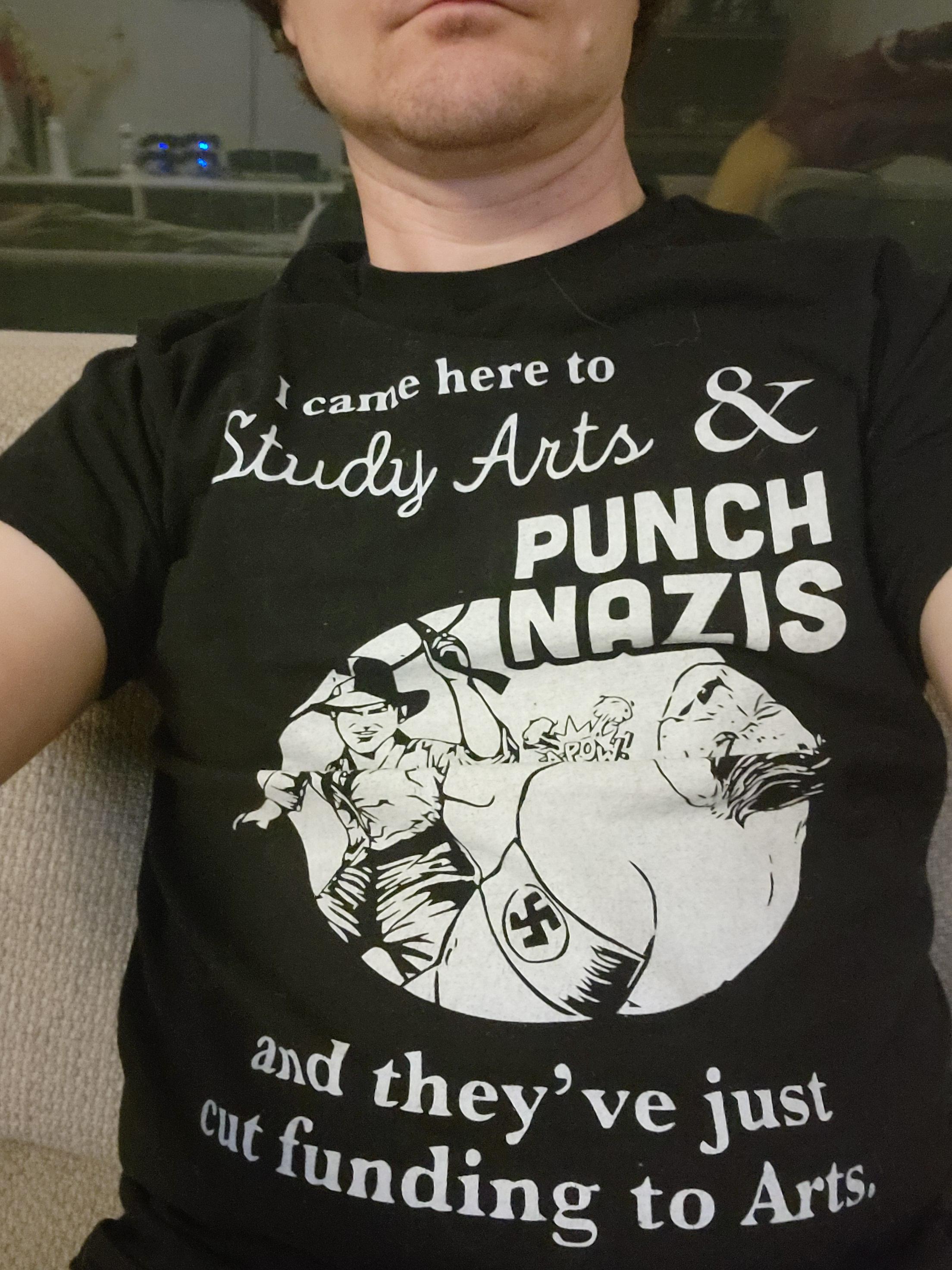 Person wearing a black shirt with white lettering and artwork. The artwork show Indiana Jones punching a Nazi. The lettering says "I came here to study arts and punch Nazis and they've just cut funding to the Arts."