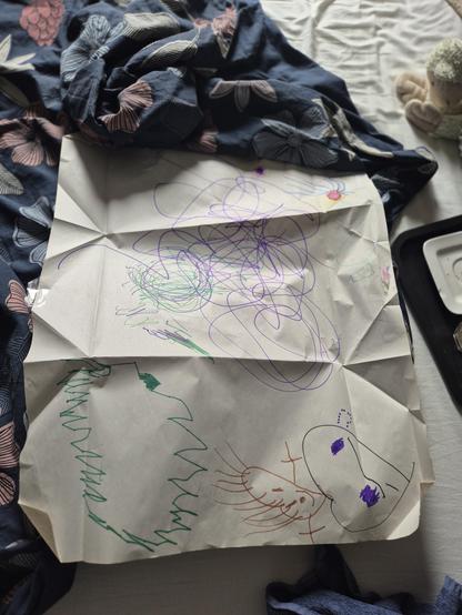Foto van bovenaf. Een groot stuk opengevouwen papier, betekend met vilstift (hartjes met spriethaar en takkenarmpjes, een grote groene getande ovaal ('bliksemschicht'), ronde paarse en groene krascirkels en het rode kernbegin van een zonnetje aan de rand). Op een bed, lege dekbedhoes in donkerblauw met bloemen. Dienblad met schotel en hoofdje en handjes van knuffelaap zichtbaar.