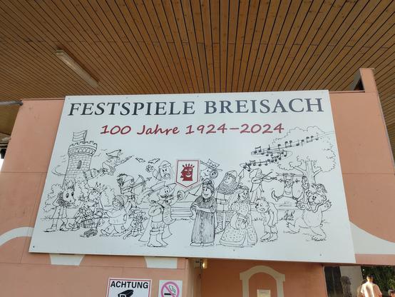 Gezeichnetes Bild zu 100 Jahre Festspiele Breisach, hängt auf der Rückseite der Sitzplatztribüne. Zu sehen sind Theatergestalten und in der Mitte in rot das Logo. Die Festspiele gibt es seit 1924.