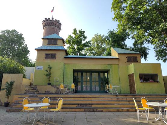 Blick auf die Bühne/Freilichtkulisse. Im Vordergrund Tische und Stühle, dann eine Treppe, dahinter ein Gebäude, was die Pension Schöller bzw. das Café darstellt. Im Hintergrund Bäume und Schlossturm.