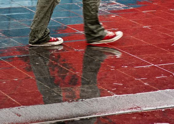 Two feet (with legs up to the knee) in brown pants and red sneakers on a wet tiled floor that is completely red except for the upper left corner. The upper left corner is blue. From the lower left to the lower right runs a metal gutter diagonally that is gleaming white in the sun.