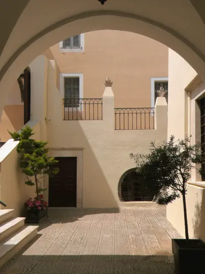 A photo of an inner courtyard at a Santorinian monastery. In the left are stairs, there are two plants to the left and right. A diagonal shadow cuts the back wall with two balustrades shielding descending (not visible) stairs going down. On the top, part of a arched ceiling is seen. The image is mostly pastel colours with green splats from the trees.