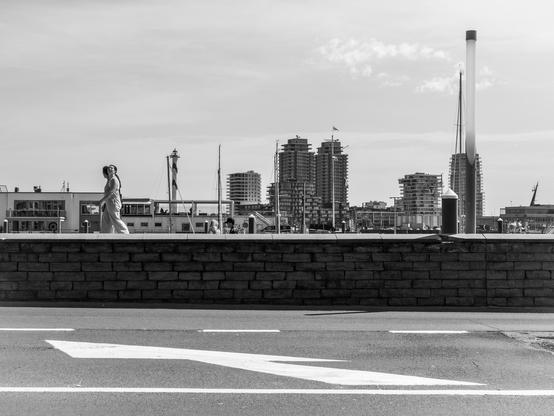 The image is a black and white photograph depicting a cityscape with a waterfront. In the foreground, a low stone wall runs horizontally across the image, with a pedestrian walking along it. The pedestrian is wearing a light-colored dress and appears to be moving towards the right side of the frame. The road in the foreground has a white arrow painted on it, indicating the direction of traffic.

In the background, a variety of buildings are visible, including high-rise residential structures and a prominent tall, slender tower with a flag at the top. Several masts, likely from boats, are visible, suggesting a marina or dock area. The sky is partly cloudy, and the overall atmosphere is calm and serene. The image captures a moment of everyday life in an urban setting, with the juxtaposition of the pedestrian and the city skyline creating a sense of scale and perspective.