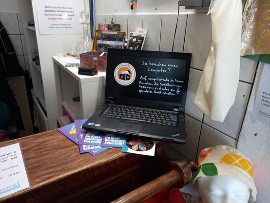 Ein alter Laptop auf einem Tresen. Auf dem schwarzen Bildschirm klebt ein Computertruhesticker, daneben wurde mit weißem Filzstift geschrieben: „Sie brauchen einen Computer? Auf computertruhe.de können Menschen, die Sozialleistungen beziehen, kostenlos ein gespendetes Gerät erhalten.“