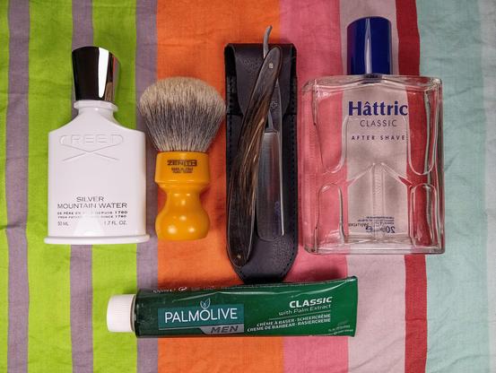 (Le Chat Mistral description)

The image displays a collection of grooming items arranged on a striped background. On the left, there is a white bottle of Creed Silver Mountain Water cologne. Next to it, a yellow-handled shaving brush with grey bristles is visible. In the centre, there is a straight razor with a wooden handle in a black leather case. To the right of the razor, there is a clear bottle of Hâttric Classic After Shave. At the bottom, there is a green tube of Palmolive Classic shaving cream with palm extract. The items are neatly laid out, suggesting a traditional shaving setup.