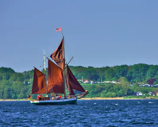 Ein Segelschiff mit zwei Masten segelt vor der grünen dänischen Küste. Die braunen Segel sind gesetzt, der Himmel ist wolkenlos. 
Der Rumpf des Schiffes ist grün, darüber die Bordwand ist weiß. Es befinden sich viele Personen an Bord, ein Teil davon ist damit beschäftigt, die Segel ordentlich in den Wind zu setzen.