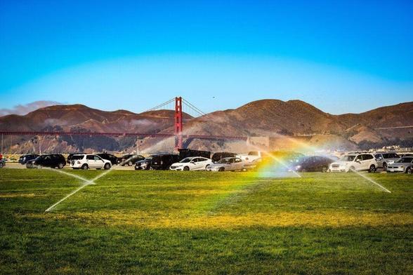 A vibrant park scene featuring a parking lot with numerous cars, primarily white and black, extending towards fog-covered hills. In the foreground is a grassy field marked with white corner lines, displaying a rainbow effect near the center. In the distant background, the iconic red structure of the Golden Gate Bridge spans the scene under a blue sky.