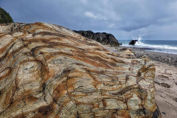 A close-up shot of a large, multi-coloured rock with intricate swirling patterns of brown, orange, and grey, dominating the foreground of a sandy beach. In the background, under a dramatic cloudy sky, waves crash against the shore and distant rocky outcrops are visible.