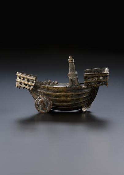 A intricately crafted incense storage container shaped like ship, made of bronze with a weathered patina. The container features a central lighthouse-like structure rising from its deck and is supported by Roman coins serving as decorative wheels, on is missing. The ship’s bow and stern are elevated and adorned with geometric carvings and porthole-like cutouts, enhancing its detailed, miniature design.